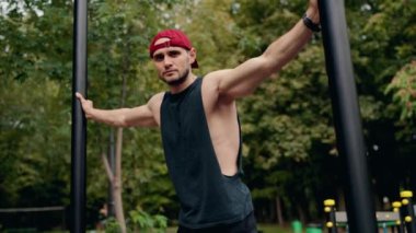 Formda bir adam ormanda bir spor salonunda kollarını parmaklıklara uzatıyor. Siyah bir atlet, koyu şort ve kırmızı beyzbol şapkası giyiyor. Esniyor, duruyor ve kameraya bakıyor..