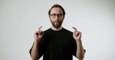Bir adam elini kaldırır ve parmaklarını bir araya getirir, bu da küçük bir boşluk ya da nesne olduğunu gösterir. Gözlük ve siyah bir tişört giyer, minimalist beyaz bir arka plana yaslanır.