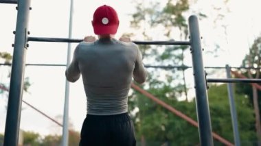 Gri tişörtlü ve kırmızı şapkalı genç bir atletik gün boyunca bir parkta yatay bir çubukta barfiks çekiyor. Sırtından vurulmuş. Fitness ve dışarıda çalışma konsepti.