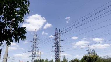 Yüksek voltajlı elektrik hatları, ağaçlarla çevrili dağınık bulutlarla açık mavi bir gökyüzü boyunca uzanıyor. Görüntü, insan yapımı altyapı ve doğal elementlerin kesişimlerini yakalıyor..