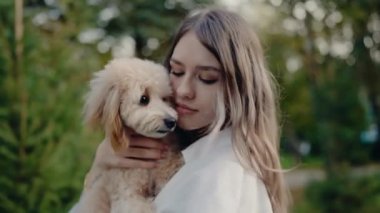 Genç bir kadın sevimli pofuduk köpeğini sakin bir park ortamında nazikçe tutuyor. Aralarındaki bağ, sakin ifadelerinde ve nazik dokunuşlarında belli oluyor..