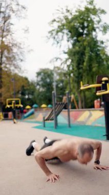Kaslı bir adam açık hava spor alanında geğirir. Tüm vücut egzersizi için şınav ve zıplama hareketlerini birleştiriyor. Kentsel ortam bu fitness rutini için bir zemin sağlıyor,