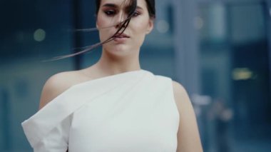 Kendine güvenen beyaz elbiseli genç bir kadın modern bir binanın önünde duruyor. O, kameraya doğru bakarken saçı rüzgarda hafifçe esiyor. Kırmızı oje ve şık bir elbise çok şık.