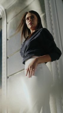 Koyu lacivert bluzlu ve beyaz etekli kendine güvenen bir iş kadını dekoratif pervazlı bir binaya karşı duruyor. Elini kalçasında gezdiriyor, profesyonellik ve stil fışkırtıyor.