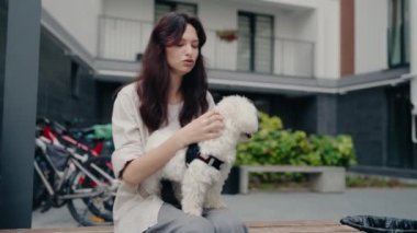 Genç bir kadın bir apartmanın önündeki bankta otururken küçük beyaz köpeğini nazikçe okşuyor. Köpek bir koşum takımı takıyor. Sahne aşkı ve özeni çağrıştırıyor.,