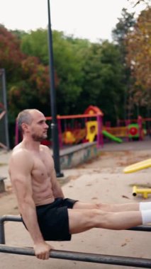 Kaslı bir adam açık hava parkı ortamında paralel barlara dalış yapar. Uygun şekle odaklanır, güç ve zindelik gösterir. Arkaplanda ağaçlar ve bir oyun alanı yer alıyor,