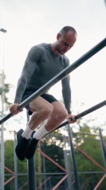 Kararlı bir adam açık hava spor salonunda paralel barlarda egzersiz yapıyor. Gücünü ve uygunluğunu gösteriyor. Dibe vuruyor, kaslarını çalıştırıyor ve sağlıklı bir yaşam tarzına bağlılığını gösteriyor..