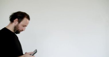 Sakallı bir adam telefonunu kullanırken durur ve onaylamayan bir bakışla kameraya bakar. Siyah bir gömlek giyiyor ve beyaz bir arka planda izole edilmiş..