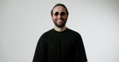 Çapraz kollu ve güneş gözlüklü gülümseyen adam. Mutlu, kendinden emin ve şık, minimalist beyaz bir zemine pozitif enerji yayıyor. Bu çok yönlü görüntü yaşam tarzı ve moda için idealdir.,