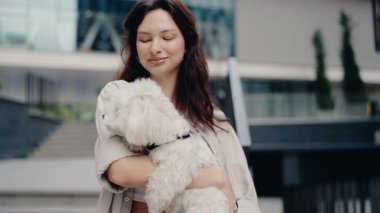Genç bir kadın küçük beyaz köpeğini kollarında tutuyor ve gülümsüyor. Modern bir binanın önündeler. Köpek bir tasma takıyor. Mutlu Genç Kadın Küçük Beyaz Köpeğini Tutuyor