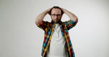 Genç bir adam hayal kırıklığı içinde ellerini saçlarında gezdiriyor. Renkli bir ekose gömlek ve gözlük takıyor beyaz bir stüdyo arka planına karşı. Bu video stres, endişe ve endişeyi betimliyor.,