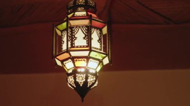 Tavandan sarkan süslü Fas feneri. Geleneksel renkli cam lamba, metal çerçeve. Ilık ışık, karmaşık Fas tasarımını aydınlatıyor. Seyahat, kültür veya ev dekorasyonu projeleri için ideal.