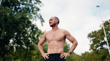 Üstsüz bir adam açık hava antrenmanı sırasında ara veriyor, elleri kalçasında, soluklanıyor. Kararlı ve fitness hedeflerine odaklanmış görünüyor. Parktaki ağaçlar doğal bir zemin sağlar..