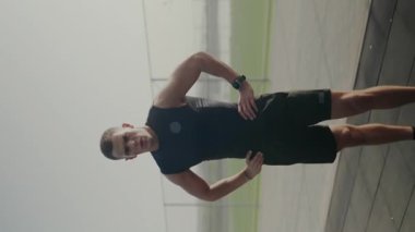 Spor kıyafetli odaklanmış genç bir adam duvara yaslanıyor, kararlılık ve güç gösteriyor. Uyanık bir atlet, teknoloji meraklısı bir sporcuyu ön plana çıkarıyor. Dikey video.