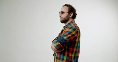 Ekose gömlekli ve güneş gözlüklü kendine güvenen bir adam kollarını kavuşturmuş, başka tarafa bakıyor ve sonra yukarı bakıyor. Tertemiz beyaz bir arka plana karşı. Yaşam tarzı, moda ve güven temaları için mükemmel.
