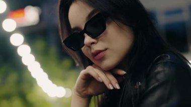 Şık bir kadın portresi güneş gözlüğü ve deri ceket takıyor, kentin gece ayininde Bokeh ışıklarıyla güven içinde poz veriyor. Güneş gözlüğü ve deri ceket giyen şık bir kadın.