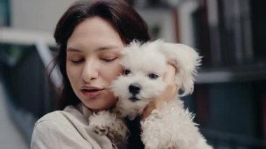 Genç bir kadın küçük, tüylü beyaz köpeğini nazikçe tutuyor. Evcil hayvanını sevgi ve özenle kucaklıyor. Köpek sakin ve merakla kameraya bakar. Aralarında özel bir bağ var..