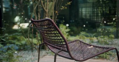 Yalnız bir hasır sandalye huzurlu bir bahçede duruyor. Yeşil yapraklar ve modern mimari rahatlamak için ideal sakin bir sahne yaratıyor. Boş Hasır Sandalye Serene Açık Bahçe Ayarları