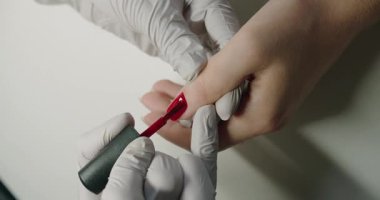 Tırnak bakımı tedavisi. Manikürcü müşteri tırnaklarını parlak kırmızı ojeyle boyuyor. Manikür ustası eldivenler içinde müşterilerinin tırnaklarına kırmızı jel sürerek güzellik salonunu boyuyor. Kadın elleri için profesyonel bakım.