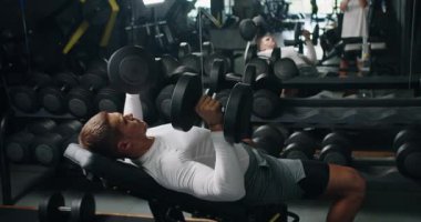 Kendini adamış genç bir adam ağır dambıllarla göğüs egzersizleri yapıyor, modern bir spor salonundaki bir bankta uzanıyor, güç ve zindelik sergiliyor. Spor salonunda Dambıllarla Odaklanmış Genç Adam Eğitim Sandığı