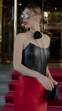 Deri bluz ve kırmızı etek giyen, güneş gözlüklü aksesuar takan, kırmızı halıda güvenle yürüyen şık bir kadın. Siyah Deri Korse Giyen Moda Kadını ve Kırmızı Etekli Şık Mekan