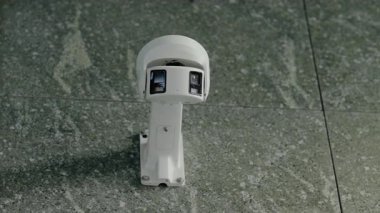 Güvenlik kamerası duvara monte edilmiş. Kamera beyaz ve iki lensi var. Beyaz metal bir desteğe bağlı. Duvar gri granit fayanslardan yapılmıştır. Bu video güvenlik duygusunu çağrıştırıyor.,