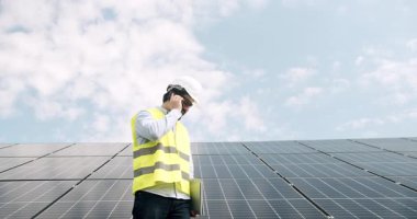 Erkek mühendis, güneş çiftliğinde akıllı telefondan konuşuyor. Düşük açılı üniformalı adam güneş enerjisi istasyonunda güneşli bir günde fotovoltaik panellerin yanında yürüyor ve bulutlu mavi gökyüzüne yapılan çağrıya cevap veriyor..