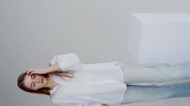 Beyaz düğmeli gömlekli genç bir kadın model, sakin ve rahat bir hava vererek tarafsız bir arkaplana karşı zarif bir şekilde poz veriyor. Beyaz Gömlekli Genç Kadın Minimalist Bir Ayarda Poz Veriyor. Dikey video.