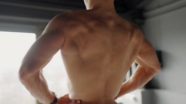 Formda, kaslı bir adamın spor salonunda durup yumuşak ışıklandırılmış arka plana karşı tonlu sırt kaslarını sergilemesi. Spor salonunda Sırtına Odaklanan Kaslı Adam