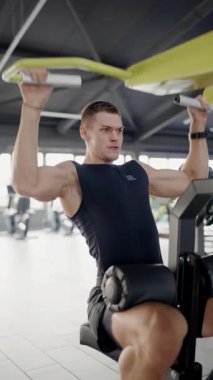 Odaklanmış genç bir adam spor salonunda yoğun bir şekilde antrenman yapıyor. Egzersiz sırasında çaba ve konsantrasyon gösteriyor. Onun ifadesi eğitiminin yoğunluğunu vurgular. Genç adam spor salonunda çok sıkı çalışıyor. Dikey