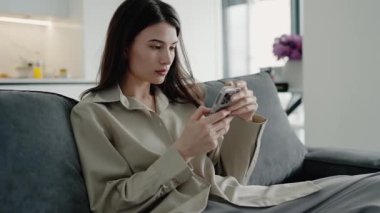 Evdeki kanepede oturan genç bir kadın, akıllı telefonunu kullanıyor. Rahatlamış ve modern oturma odasındaki mobil cihazına odaklanmış..