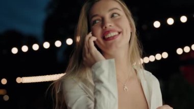 Beyaz bluzlu neşeli bir genç kadın, arka planda sicim ışıkları olan bir gece partisinde cep telefonuyla konuşurken gülümsüyor. Gece partisinde telefonda konuşurken gülümseyen genç kadın.