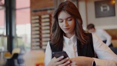 Odaklanmış genç bir kadın akıllı telefonunu iyi ışıklandırılmış, modern bir kafede otururken kullanır, muhtemelen bir mola sırasında ya da sıradan bir iş toplantısında. Moda Kafe 'de Akıllı Telefon kullanan Genç Profesyonel Kadın