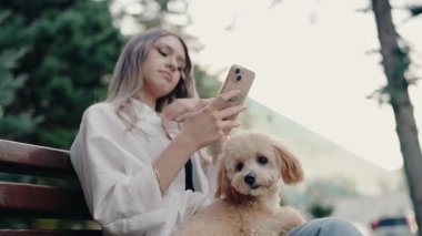Genç bir kadın sevimli tüylü köpeğiyle bankta oturuyor ve telefonunu kullanıyor. O, bakınırken nazikçe köpeğini okşar. Köpek sakince oturur. Sakin bir şehir ortamında eğlence, teknoloji ve evcil hayvanlar..