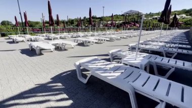 Temiz mavi gökyüzünün altında düzgün düzenlenmiş beyaz şezlonglar ve kapalı şemsiyelerin yer aldığı boş bir tatil köyü manzarası. Tatil, rahatlama ve güneş banyosu için mükemmel..