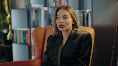 Ofis koltuğunda oturan siyah takım elbiseli genç bir iş kadını düşünceli ve düşünceli görünüyor. Arkaplandaki kitaplıklar profesyonel bir dokunuş ekliyor. Genç Güzel Kadın