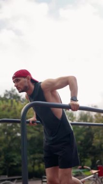 Kararlı bir erkek sporcu, açık hava parkı ortamında paralel barlara daldı. Egzersizine odaklanıyor, güç ve zindelik gösteriyor. Sahne adanmışlığın özünü yakalar.