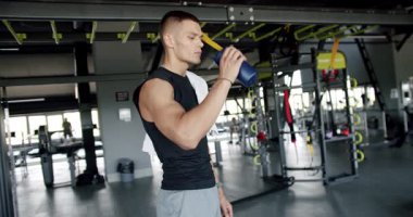 Kendini adamış genç bir adam modern bir spor salonunda egzersiz yaparken su içer. Bu görüntü bir ferahlatıcı anı yakalar. İyi donanımlı bir spor salonunda Odaklanmış Atletizm İçme Suyu