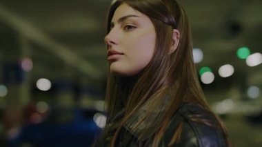 Deri ceketli kadın park halindeki arabalara karşı büyüleyici bir görsel birliktelik yarattı. Şık bir kız. Genç, lüks, seksi bir kadın karanlık arka planda kameraya bakıyor..