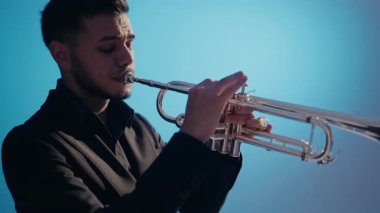 Takım elbiseli genç ve yakışıklı bir adam arzu ve ilhamla trompet çalar. Modern bir stüdyoda mavi arka planda trompet çalan bir erkek caz şarkıcısı. Canlı caz konseri..