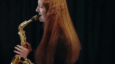 Siyah elbiseli genç bir kadın stüdyoda siyah arka planda saksafon çalıyor. Saksafon çalan bir bayan müzisyen. Siyah bir gece elbisesi giymiş, sahnede solo yapan bir kadın saksafoncu..