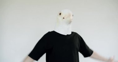 Beyaz kuş maskesi takan biri, minimalist, açık beyaz bir arkaplana karşı duruyor. Elleri ve kolları açık hareket ediyorlar, gerçeküstü ve ilgi çekici bir sahne yaratıyorlar..