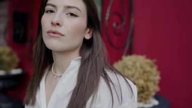 Beyaz bir bluz giymiş genç ve sofistike bir kadının canlı kırmızı bir arkaplanda durduğu, sakin ve kendinden emin bir tavır sergilediği yakın plan..