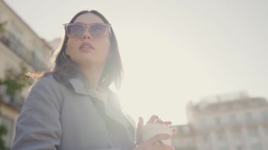 Kendine güvenen genç bir kadın güneşli şehir sokaklarında geziyor, şık bir trençkot ve güneş gözlüğü takmış bir kahvenin tadını çıkarıyor. Güneşli bir günde şehirde elinde kahveyle dolaşan zarif bir kadın.