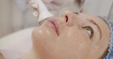 Spa 'da profesyonel yüz bakımı gören bir kadın. Güzellik merkezinde modern bir aletle rahatlatıcı bir yüz bakımı alan bir kadının yakın çekim görüntüsü..