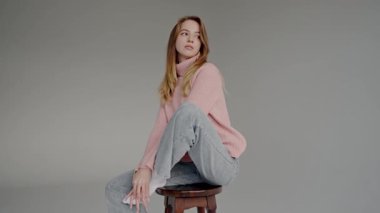 Rahat pembe bir balıkçı yaka ve rahat gri bir kot giyen zarif genç bir kadın, düşünceli bir ifadeyle huzur içinde oturuyor. Pembe Kazaklı ve Gri Kot Giyen Serene Genç Kadın