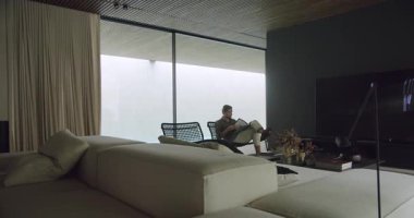 Görüntü, şık, modern bir ev ortamında uzanan sakin ve sofistike bir yaşam tarzı yayan bir adamı yakalıyor. Adam Minimalist Dekorla Modern Ev İçlerinde Rahatlıyor