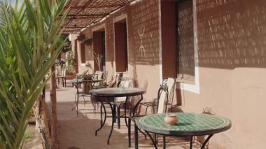 Geleneksel masaları, sandalyeleri ve gölgesi olan sakin bir Fas avlusu. Güneş ışığı sazlığın çatısından süzülüp binanın üzerine gölge düşürür. Kafe turistler için rahatlatıcı bir atmosfer sunuyor..