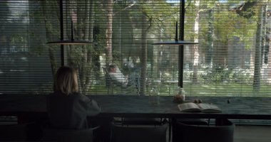 Modern, karanlık bir ahşap masaya oturmuş, dışarıdaki manzaraya dikey perdelerden dikkatlice bakan bir kadının sakin bir sahnesi. Doğal ışıkla dolu sakin bir ofis..