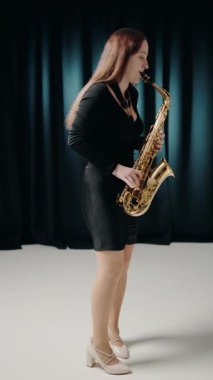 Saksafon çalan caz müzisyeni. Siyah bir gece elbisesi giyen kadın bir saksafoncu sahnede solo performans sergiliyor. Siyah elbiseli genç bir kadın saksafon çalıyor. dikey